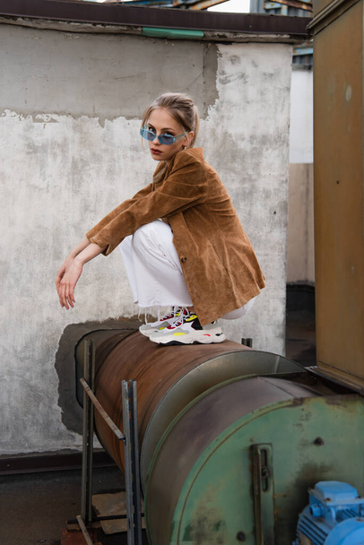 full length of young woman in stylish outfit with suede blazer and blue sunglasses posing on rusty construction  - Photo, Image