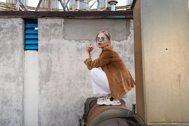 comprimento total da jovem mulher na roupa da moda e óculos de sol azuis posando na construção enferrujada  - Foto, Imagem