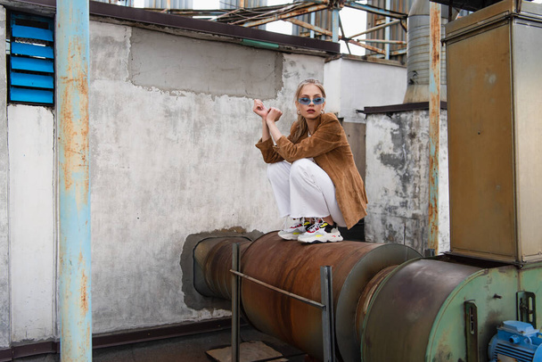 full length of young stylish woman in trendy outfit and blue sunglasses posing on rusty construction  - Foto, immagini
