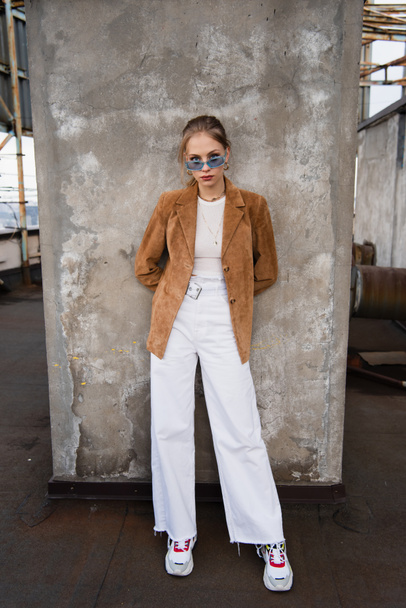 full length of young model in blue sunglasses and stylish outfit posing near concrete wall - Foto, afbeelding