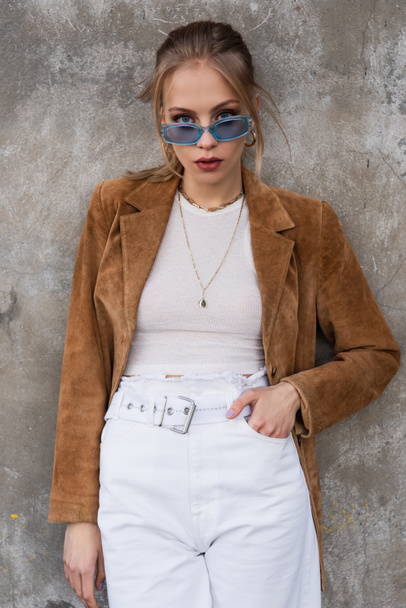 young model in blue sunglasses and stylish outfit posing with hand in pocket near concrete wall - Photo, Image
