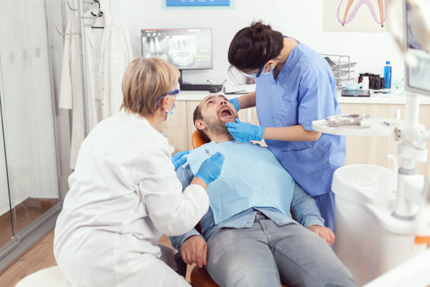 Zieke man zittend op tandartsstoel met open mond terwijl medische verpleegkundige - Foto, afbeelding