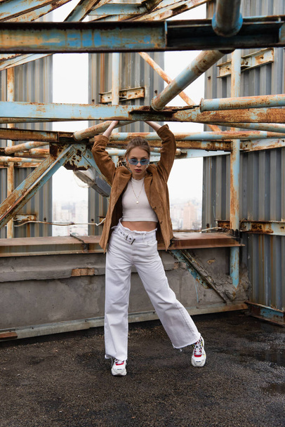 full length of pretty young model in blue sunglasses posing near weathered construction on rooftop - Photo, Image