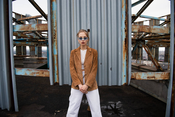 blonde model in blue sunglasses posing near rusty construction on rooftop - Zdjęcie, obraz