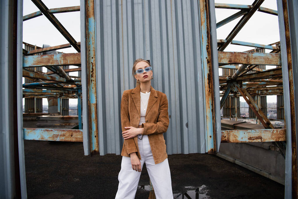 mujer bonita en gafas de sol azules y ropa elegante posando cerca de la construcción oxidada en la azotea - Foto, Imagen