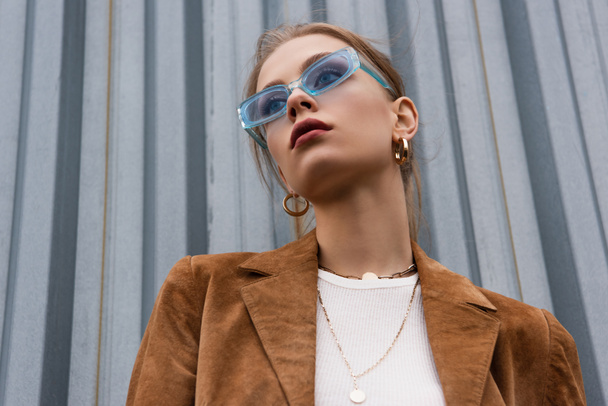 low angle view of pretty woman in blue sunglasses and stylish outfit posing outside - Fotografie, Obrázek