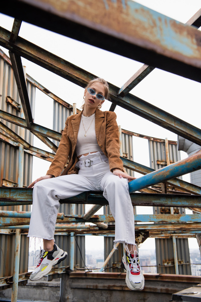 blonde woman in blue sunglasses and suede jacket posing on rusty construction of rooftop  - Fotó, kép
