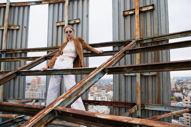 young and pretty model in stylish outfit and sunglasses standing on weathered construction of rooftop - Photo, Image