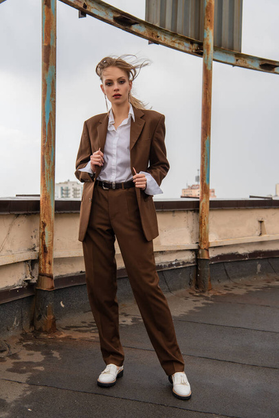 longitud completa de modelo joven en pendientes, camisa blanca y traje marrón mirando a la cámara mientras posan en la azotea  - Foto, imagen