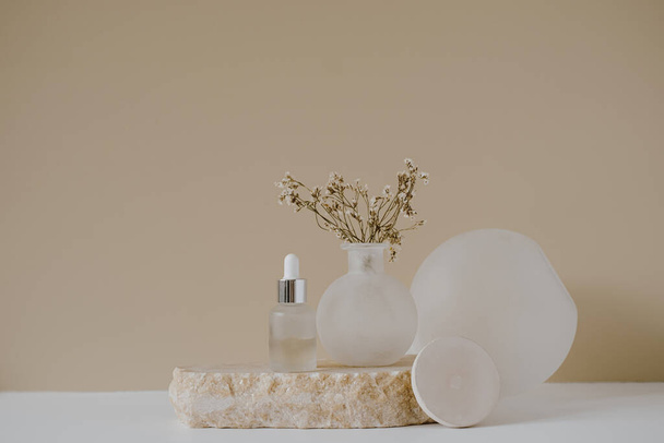 Μπουκαλάκι γυάλινο με πιπέτα, πέτρα με λουλούδια σε ουδέτερο παστέλ μπεζ φόντο. Αισθητική ελάχιστη σύνθεση ομορφιάς με περιποίηση προσώπου σώματος οργανικό φυσικό προϊόν spa - Φωτογραφία, εικόνα