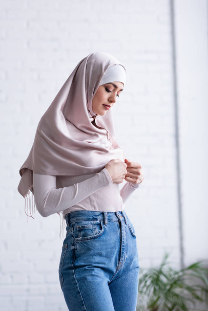 shy muslim woman touching hijab while standing at home - Фото, изображение
