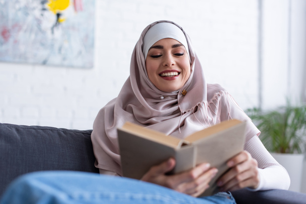 cheerful muslim woman reading book at home, blurred foreground - Фото, зображення