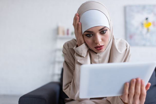 shocked muslim woman touching head while looking at digital tablet at home - Фото, зображення