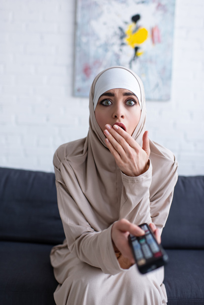 scossa donna musulmana che copre la bocca con la mano mentre guarda la tv a casa - Foto, immagini