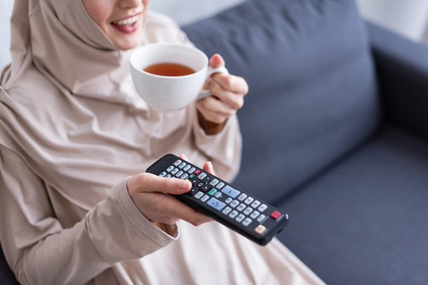 περικοπή άποψη του χαμογελώντας muslim γυναίκα με φλιτζάνι τσάι και τηλεχειριστήριο βλέποντας τηλεόραση στο σπίτι - Φωτογραφία, εικόνα