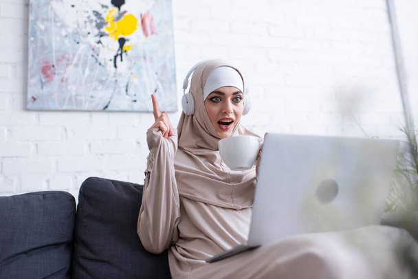 изумленная мусульманка с чашкой чая демонстрирует жест идеи во время онлайн урока - Фото, изображение