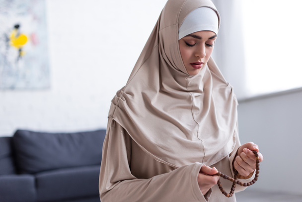 νεαρή μουσουλμάνα γυναίκα με κομπολόι προσεύχεται με κλειστά μάτια στο σπίτι - Φωτογραφία, εικόνα