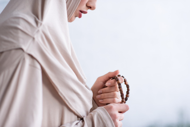 vista parcial de la joven árabe sosteniendo rosario durante la oración en casa - Foto, Imagen