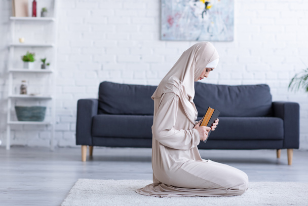 πλαϊνή άποψη των μουσουλμάνων γυναίκα προσεύχεται με κοράνι στο πάτωμα στο σπίτι - Φωτογραφία, εικόνα