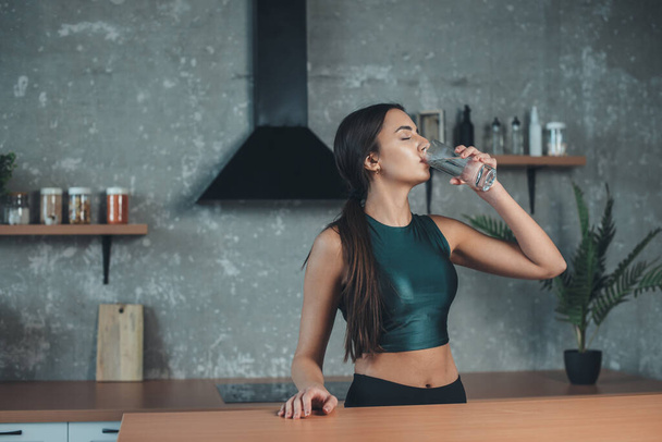 Mujer caucásica usando ropa deportiva está bebiendo un vaso de agua para hidratar su cuerpo en la cocina - Foto, Imagen