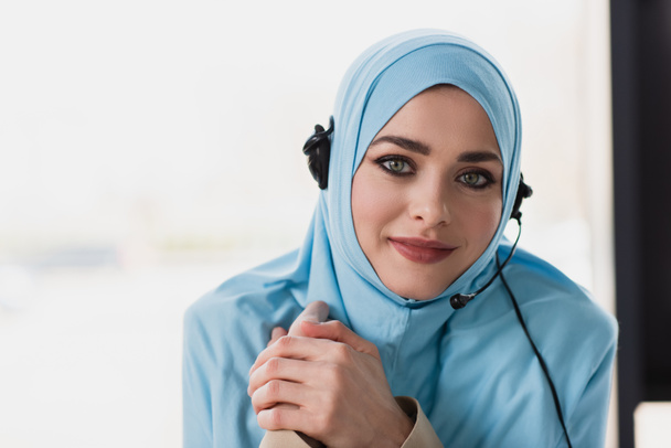 pretty muslim woman in hijab and headset smiling at camera in office - Φωτογραφία, εικόνα