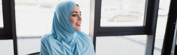 joyful muslim businesswoman looking away near window in office, banner - Foto, imagen