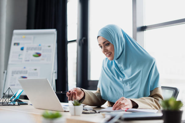 smiling muslim businesswoman pointing at laptop while working in office, blurred foreground - Photo, Image