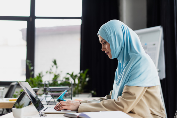 side view of young arabian businesswoman in hijab typing on laptop in office - Photo, Image