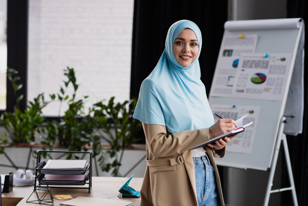 happy muslim businesswoman writing in notebook near flip chart on blurred background - Valokuva, kuva