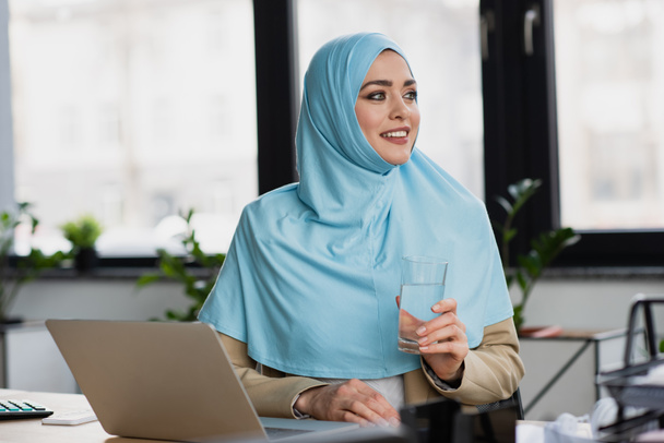 successful muslim businesswoman with glass of water looking away near laptop in office - Foto, Bild