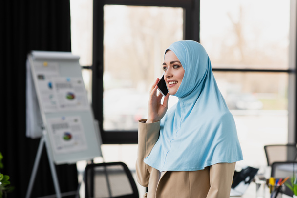 smiling muslim businesswoman talking on smartphone near blurred flip chart - Foto, imagen