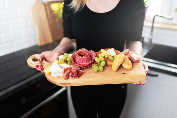 A young woman holds a serving board with food in her hands. Wooden cutting board. The kitchen is visible in the background. Square frame. There are cheese, cold cuts and fruit on a wooden board.  - Foto, Imagem