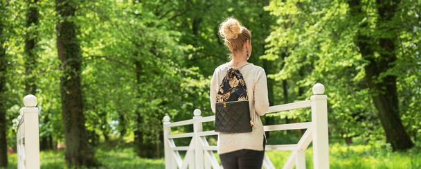 Mladá žena na túře se prochází v parku. Podívá se na zelené stromy z bílého mostu. Má na sobě batoh. Aktivní dovolená. Výlet do lesa nebo parku. Přírodní podmínky.  - Fotografie, Obrázek