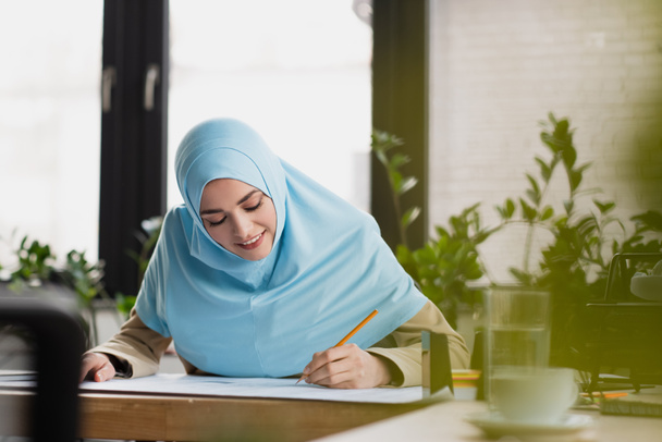 smiling arabian engineer in hijab drawing blueprint on blurred foreground - 写真・画像