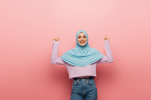 mujer musulmana emocionada en hijab y vaqueros mostrando gesto de regocijo sobre fondo rosa - Foto, imagen