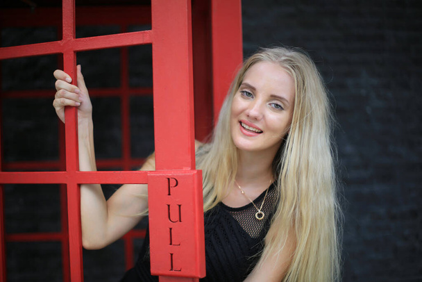 Retrato de bela menina de cabelo loira no vestido preto em pé na cabine de telefone vermelho contra a parede preta como retrato de moda pose ao ar livre. - Foto, Imagem
