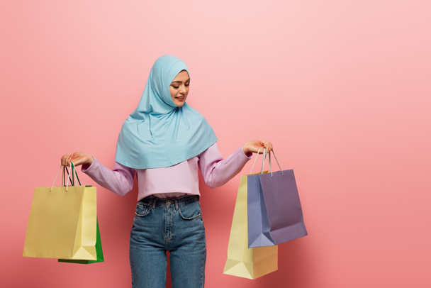 young muslim woman in hijab and jeans holding multicolored shopping bags on pink background - Photo, image