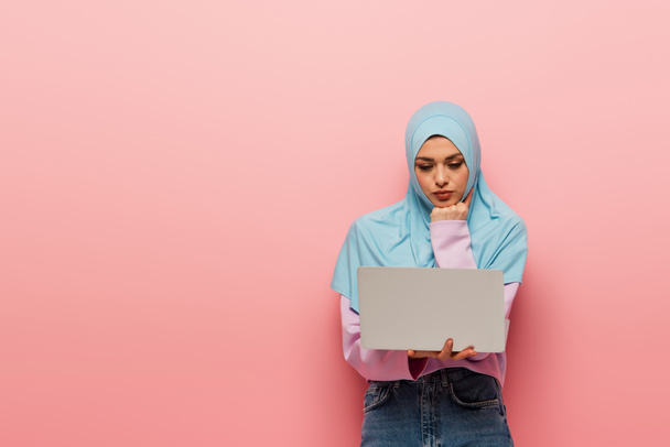 femme musulmane réfléchie regardant ordinateur portable sur fond rose - Photo, image