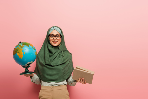 joyful muslim teacher with globe and textbooks looking at camera on pink background - Foto, imagen