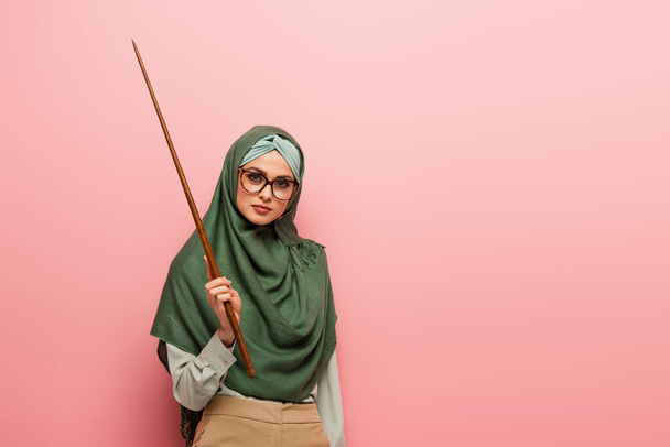young arabian teacher in green hijab holding pointing stick isolated on pink - 写真・画像