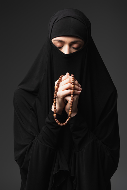 mladá muslimka se zavřenýma očima drží růženec při modlitbě izolované na černé - Fotografie, Obrázek