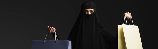 αραβική γυναίκα σε μαύρο niqab με πολύχρωμα τσάντες ψώνια που απομονώνονται σε μαύρο, banner - Φωτογραφία, εικόνα