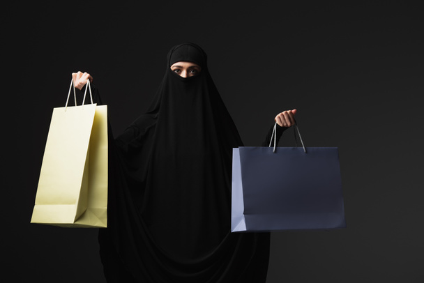 мусульманська черниця в чорному нікаб тримає різнокольорові сумки для покупок ізольовані на чорному
 - Фото, зображення