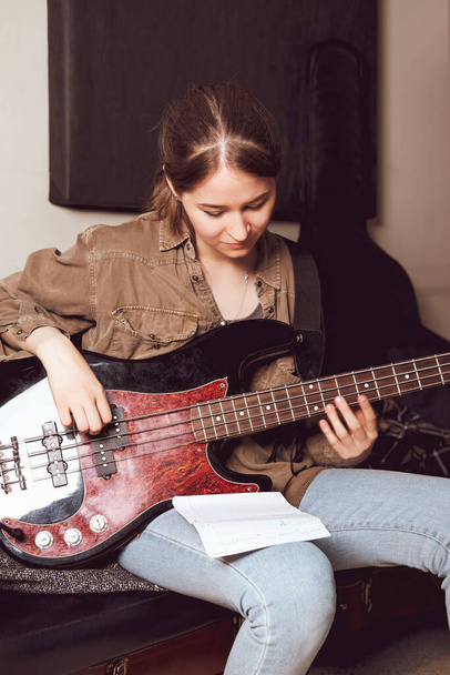 Młoda kobieta gra na gitarze basowej i patrzy na nuty akordów. sala prób. kobiecy muzyk rockowy. wielkość pionowa.  - Zdjęcie, obraz