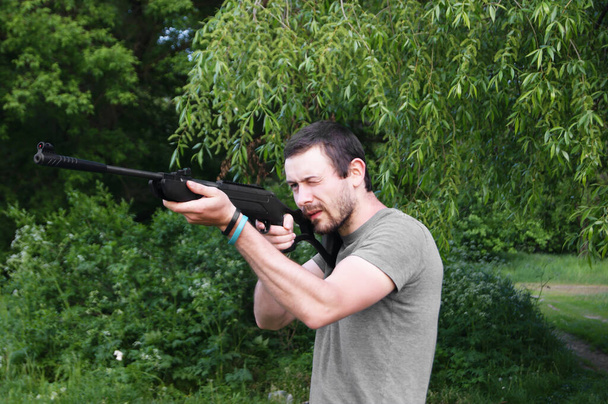Genç bir adam pnömatik bir silahtan ateş etmeye hazırlanıyor. Ağaçtan sarkan bir kavanoza nişan alır. - Fotoğraf, Görsel