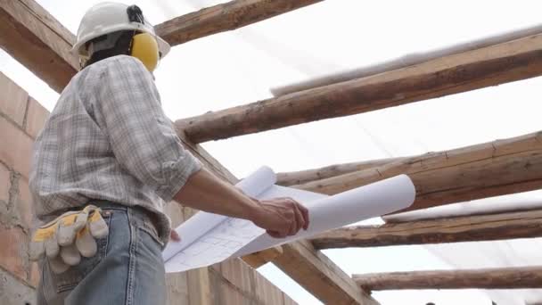 architect man at work, wearing helmet and headphones looking at blueprint , check the construction house project plan, in renovation building site background with the old wooden beams of the roof - Footage, Video