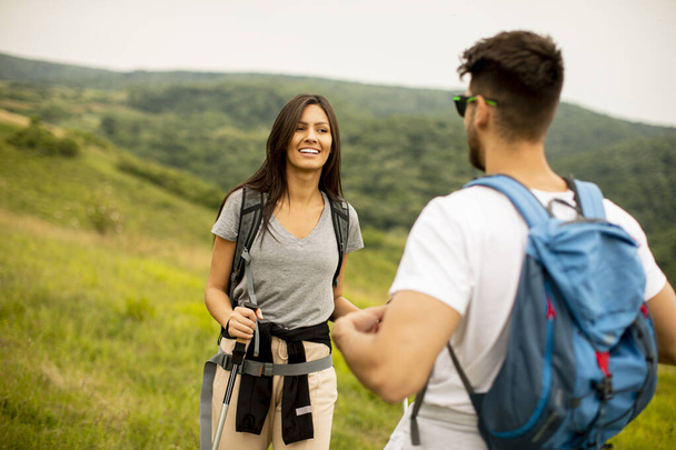 Улыбающаяся молодая пара идет с рюкзаками на зеленых холмах в летний день - Фото, изображение