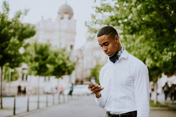 Bonito jovem empresário afro-americano usando um telefone celular enquanto espera por um táxi esperando um táxi em uma rua - Foto, Imagem