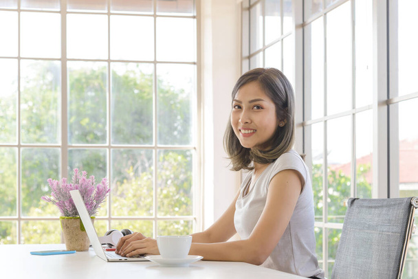 Uśmiechnięta młoda Azjatka nosi luźne ubrania pracując z laptopem w salonie w domu. Praca w domu. Patrzę w kamerę. - Zdjęcie, obraz