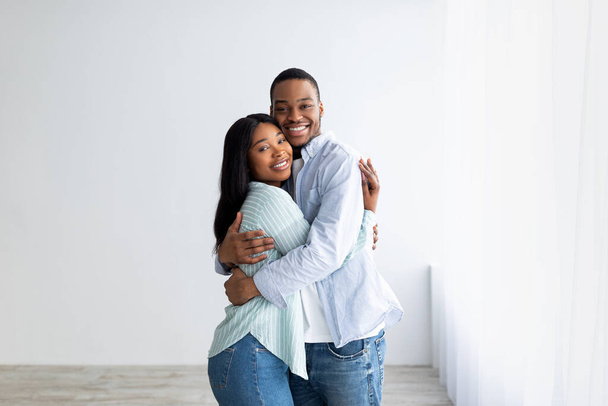 Αγαπώντας ένα ζευγάρι Αφροαμερικανών που στέκονται στο σπίτι τους, αγκαλιασμένοι και χαμογελαστοί, χαρούμενοι που αγοράζουν το δικό τους κατάλυμα - Φωτογραφία, εικόνα